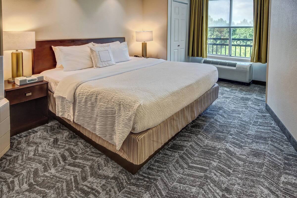 Uma cama bem arrumada com edredom branco e travesseiros, uma mesa de cabeceira com abajur, uma janela com cortinas e um radiador.