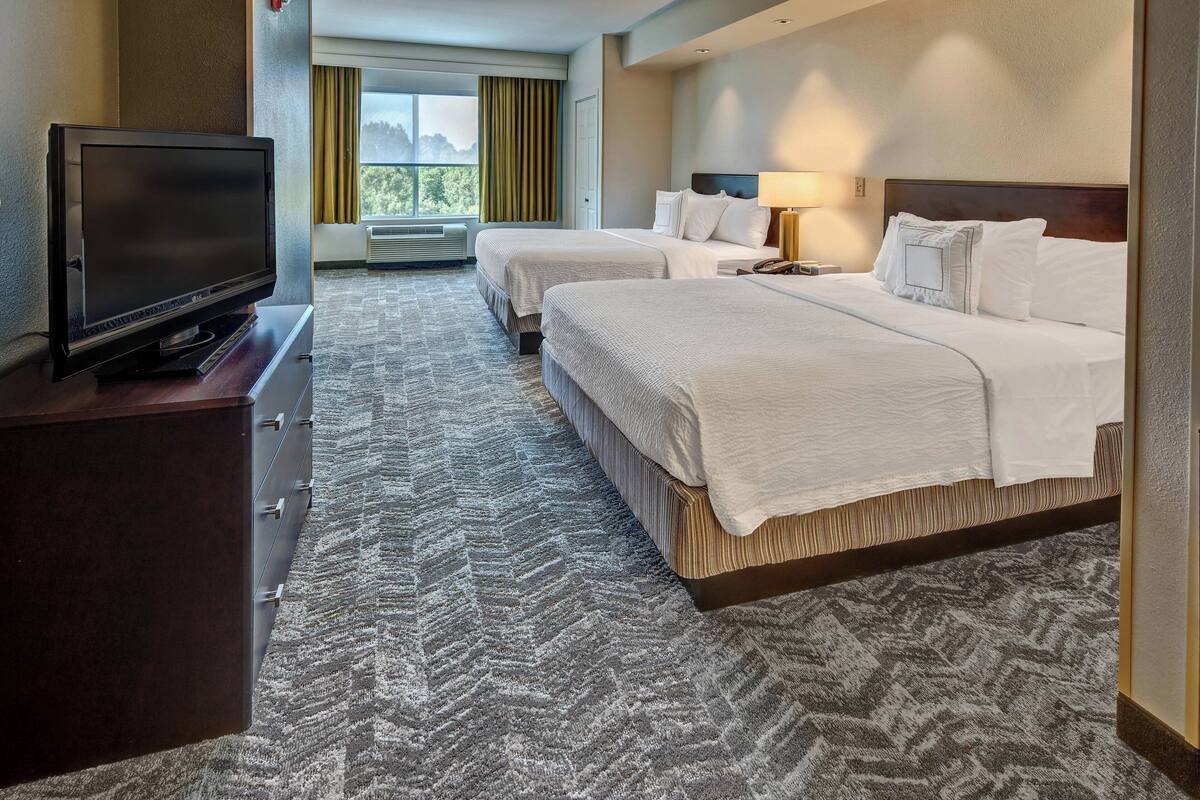 Quarto de hotel com duas camas, uma televisão em uma cômoda e uma janela grande com cortinas.