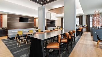 A modern hotel lobby with a large central table, seating, a TV, and a bar area.