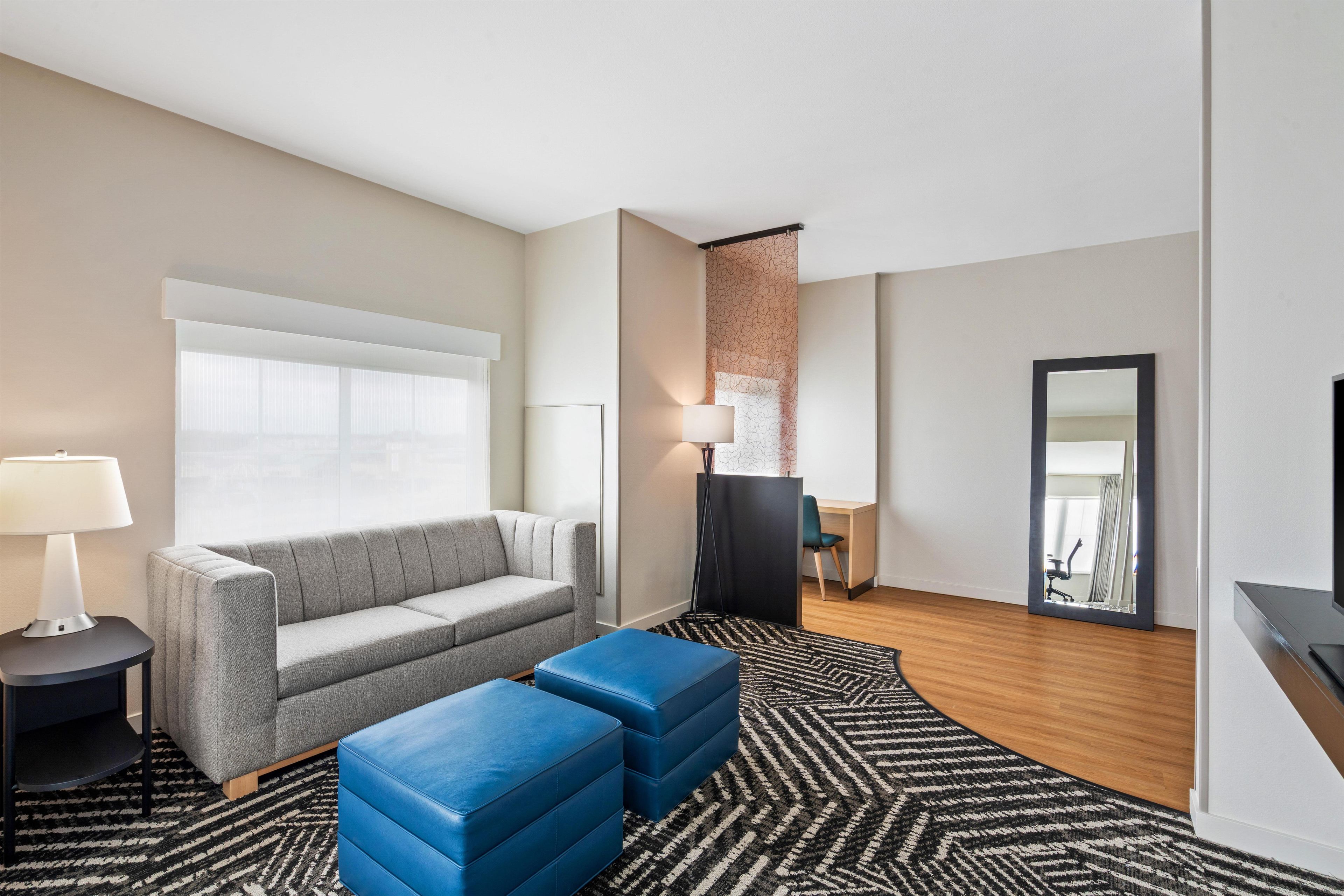 A modern hotel room with a grey sofa, blue ottomans, a black floor lamp, and a large window.