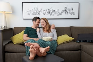 Un couple est assis sur un canapé. Au-dessus d’eux, un tableau représentant un paysage urbain est accroché au mur. Une lampe se trouve sur une table d’appoint, et un bol de pop-corn est posé à proximité.