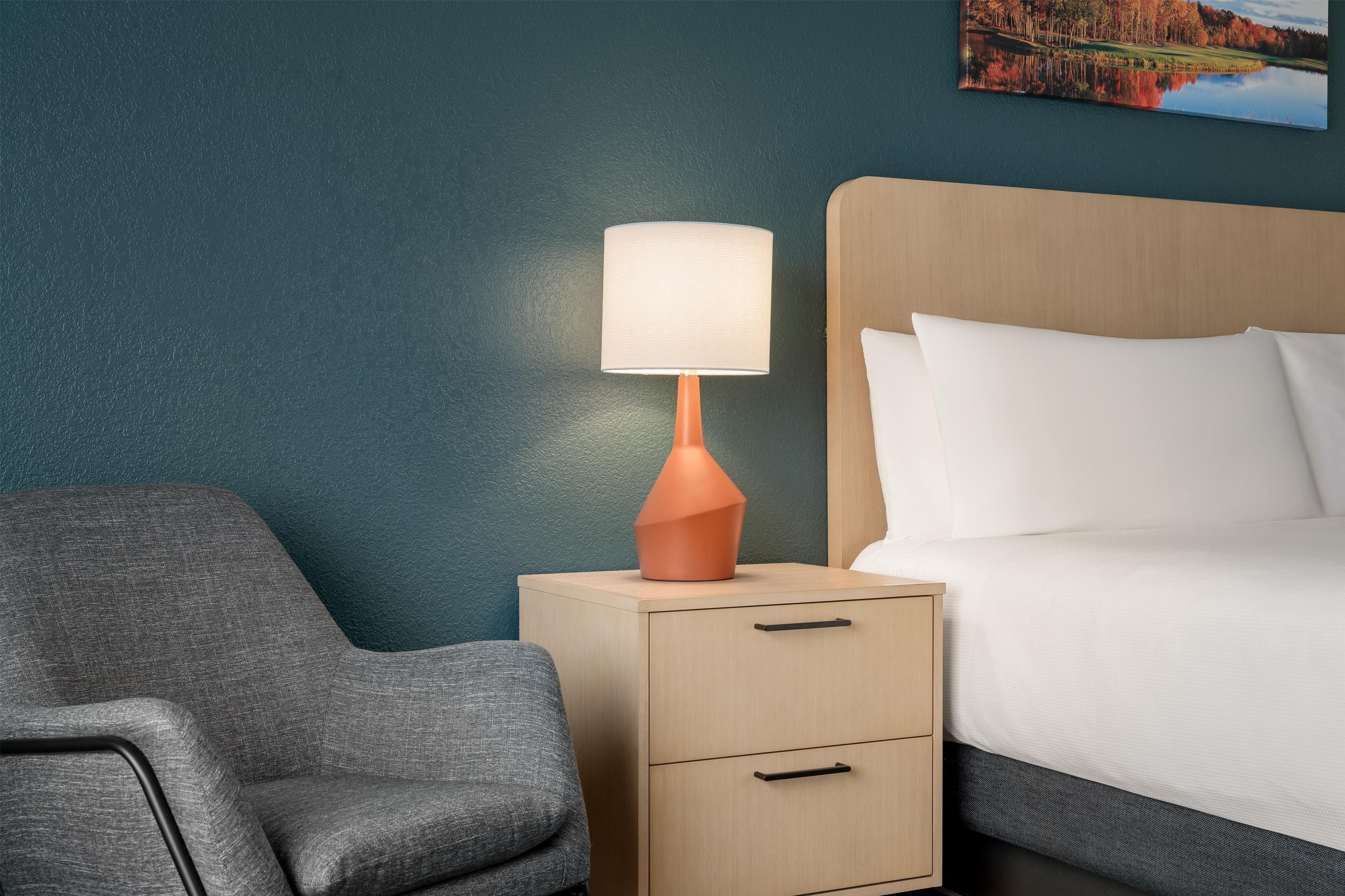 Una habitación de hotel con cama, mesita de noche con lámpara, una silla y un cuadro en la pared.