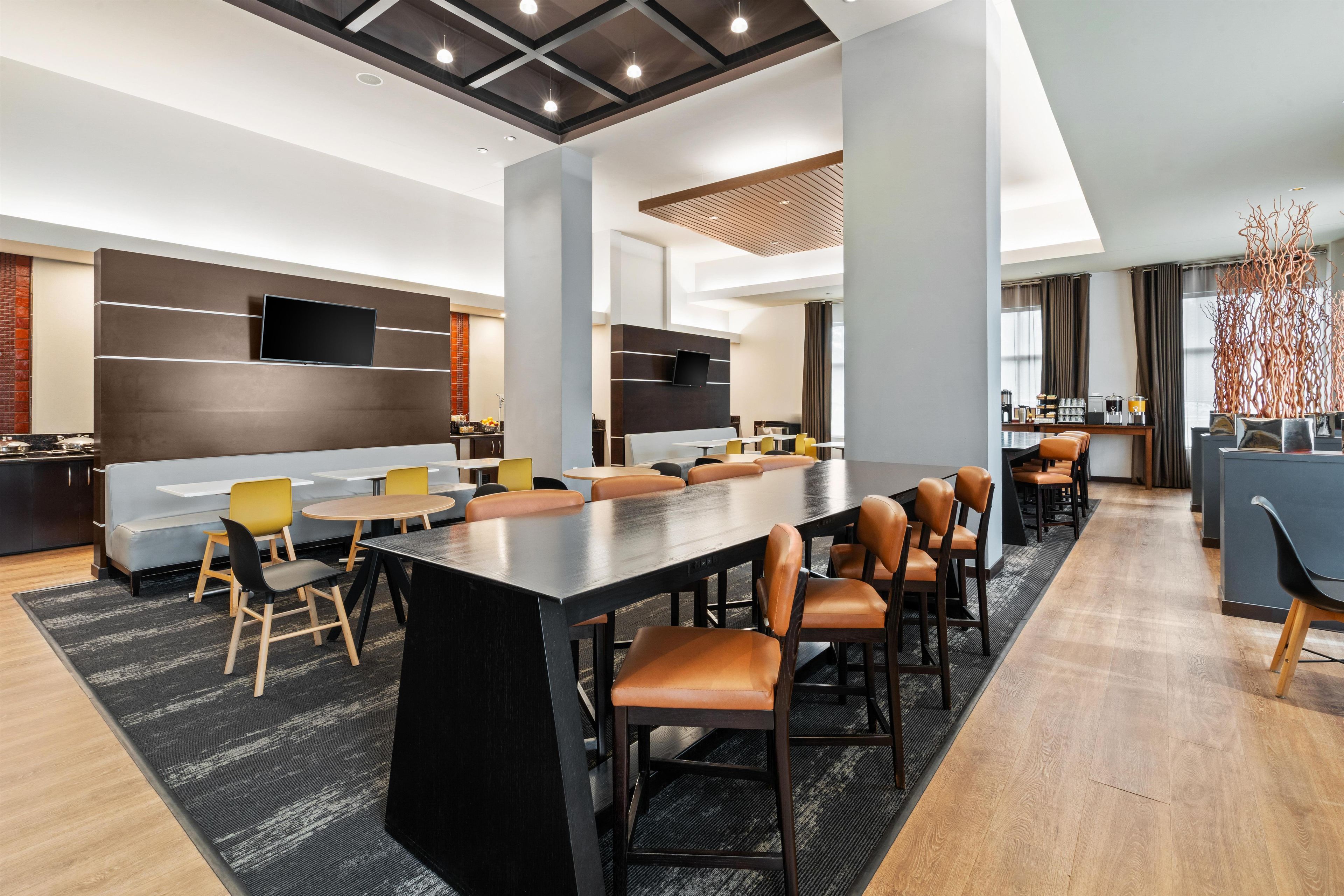 A modern hotel lobby with a large central table, seating, a TV, and a bar area.