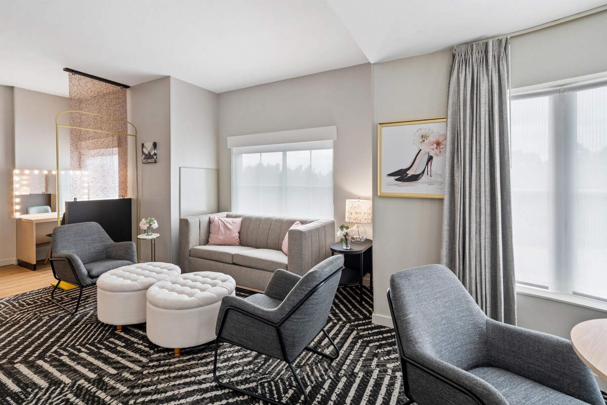 Een moderne hotelkamer met een grijze bank, twee grijze fauteuils, een ronde salontafel, een klein bijzettafeltje met een lamp, een spiegel met verlichting, een groot raam met gordijnen en een lijst met een afbeelding aan de muur.