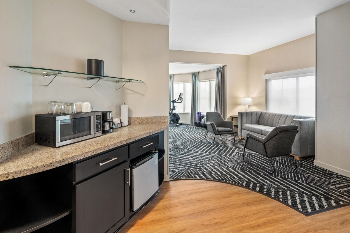 Een hotelkamer met een kitchenette, een zithoek met een bank en fauteuils, een kamerkleed met een patroon en een groot raam met gordijnen.