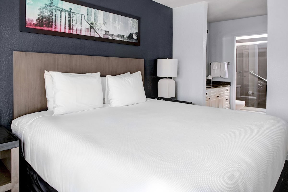 Une chambre d’hôtel avec un grand lit, une table de chevet, une lampe, un tableau encadré au mur et une vue sur la salle de bain.