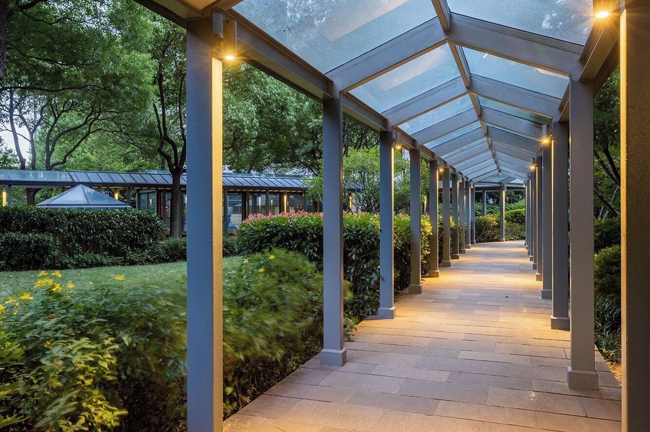 A covered walkway with glass panels and columns, surrounded by greenery and trees.