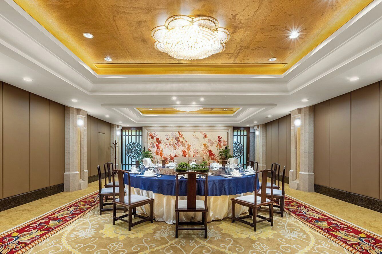 A formal dining room with a round table set for six, surrounded by chairs, with a chandelier above and a patterned carpet.