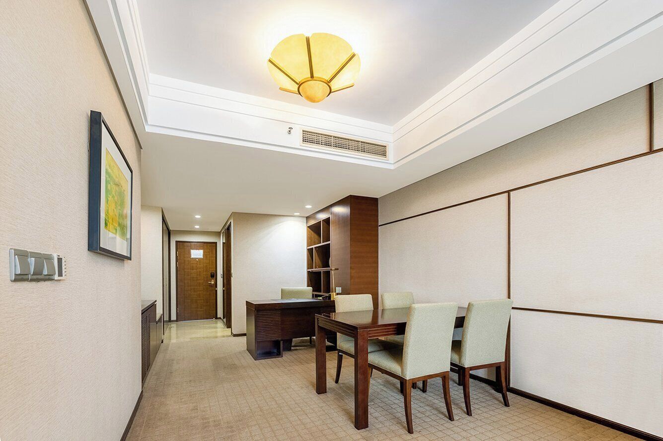 A hotel room with a dining area featuring a wooden table and chairs, a ceiling light fixture, and a door leading to another room.
