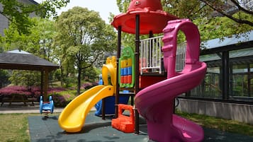 Explore a vibrant outdoor play area surrounded by lush greenery and trees, featuring a slide and climbing elements.
