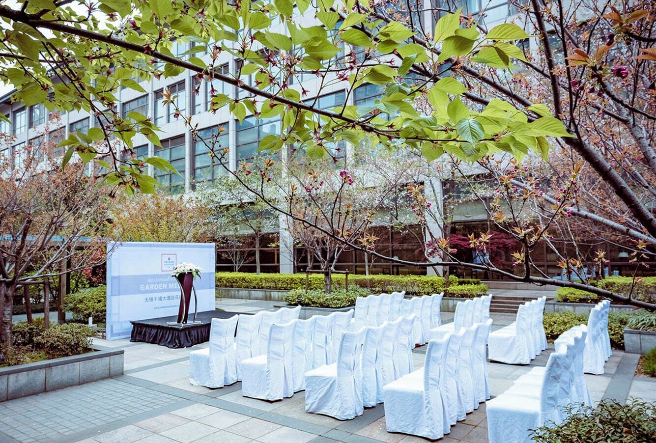An outdoor seating area with white-covered chairs arranged in rows, a stage with a speaker, and a banner in the background.