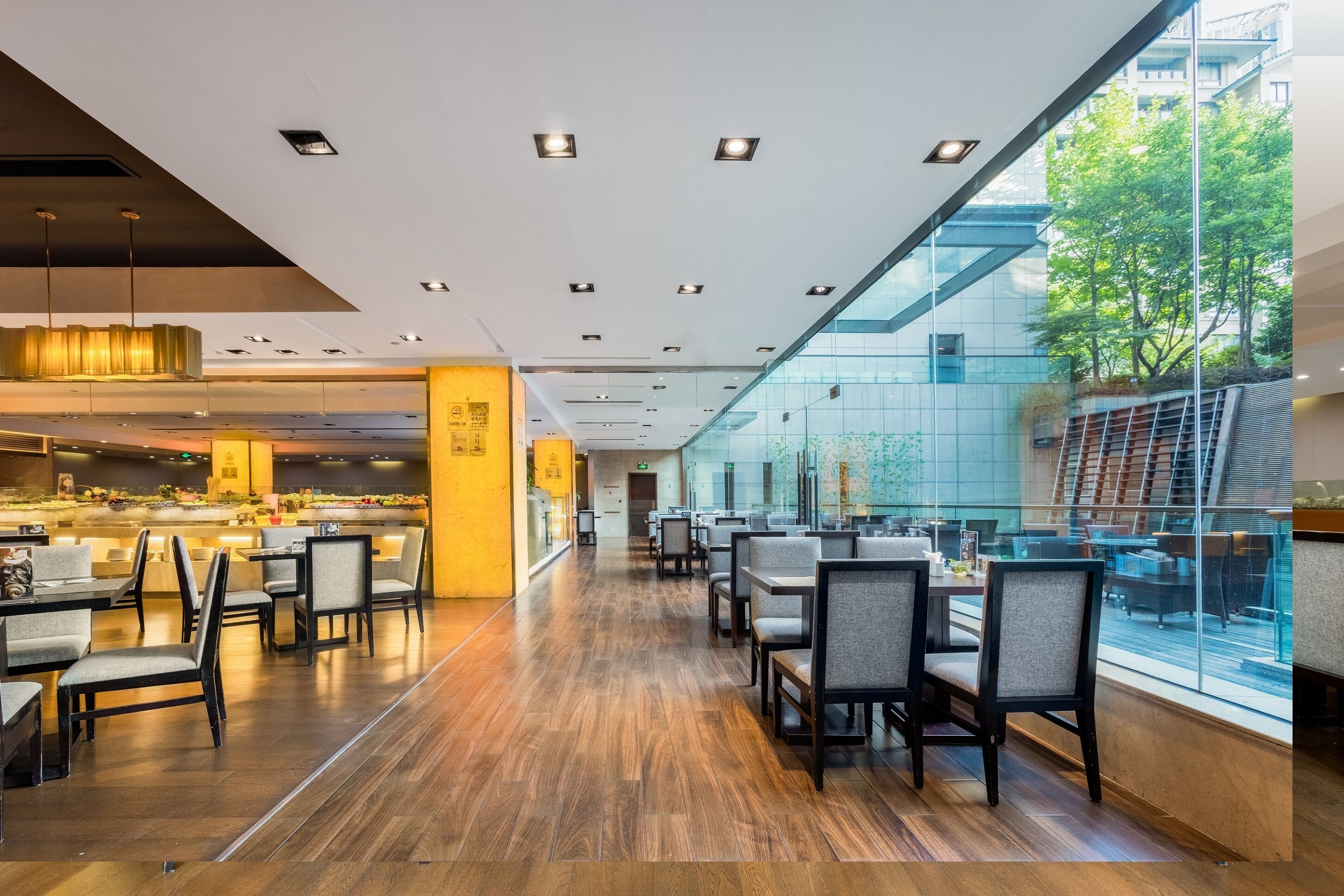 A modern restaurant with a long counter, multiple tables, and chairs. There is a glass wall on one side, and a view of trees outside.