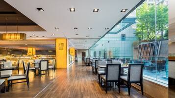 A modern restaurant with a long counter, multiple tables, and chairs. There is a glass wall on one side, and a view of trees outside.