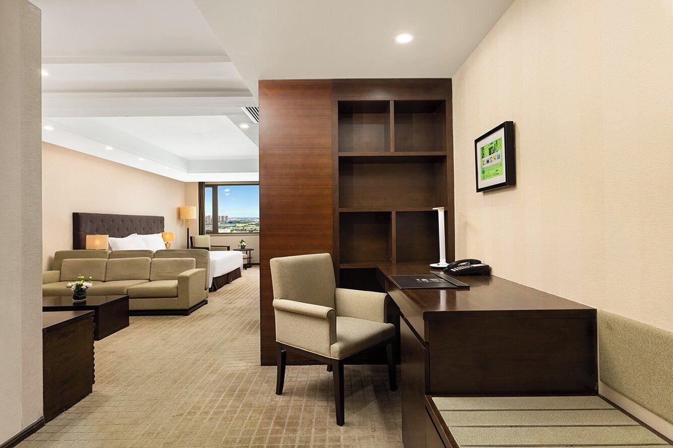 A hotel room with a bed, a sofa, a desk, and a chair. There is a wooden shelf unit and a framed picture on the wall.