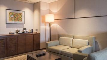 A modern hotel room with a sofa, a coffee table, a desk with a lamp, and a framed abstract painting on the wall.