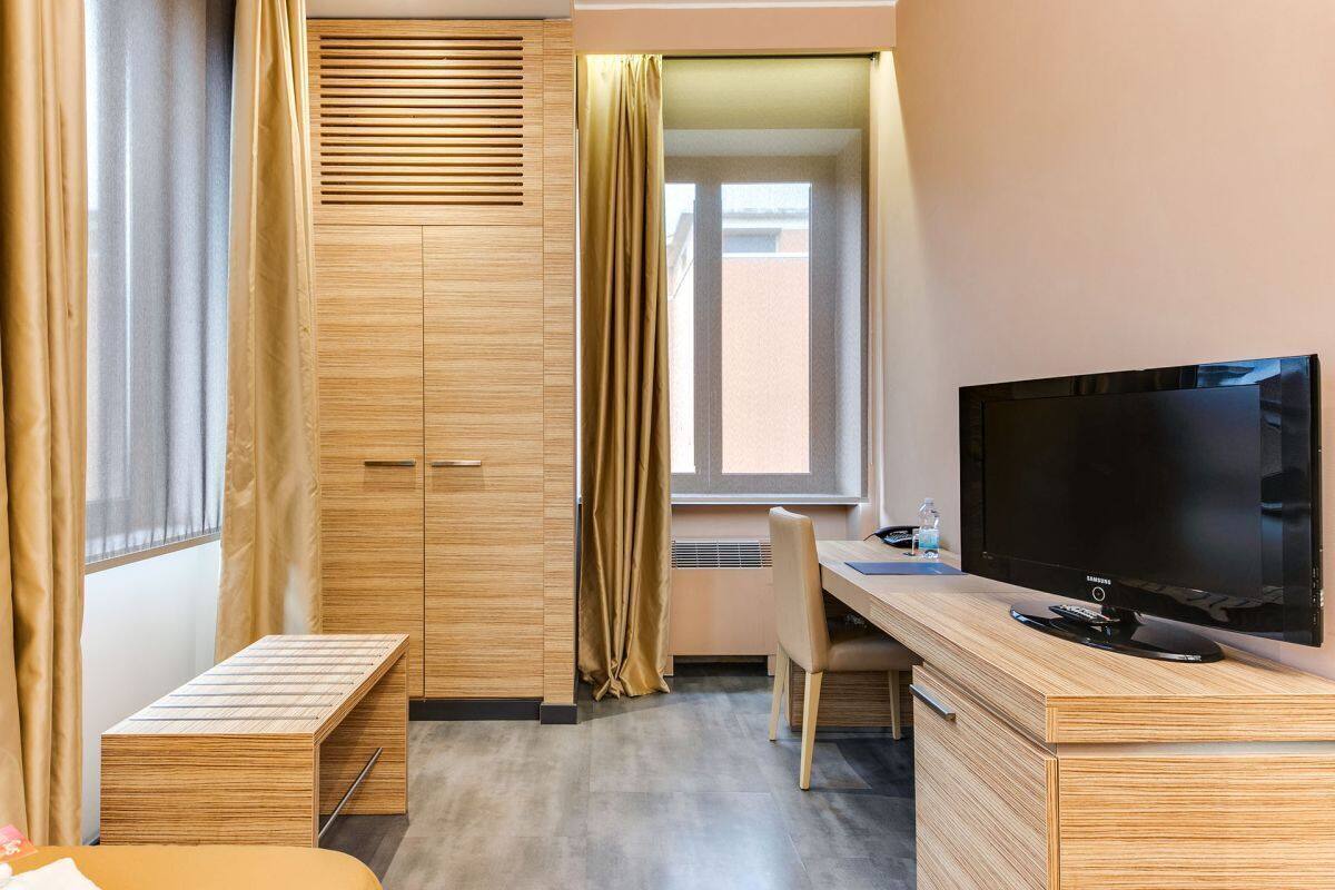 A hotel room with a wooden desk, a television, a bed, a wardrobe, and a window with curtains.