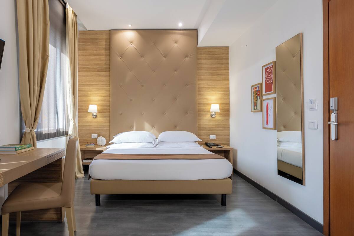 A modern hotel room with a large bed, a desk, and a chair. There are two bedside tables with lamps and a large mirror on the wall.