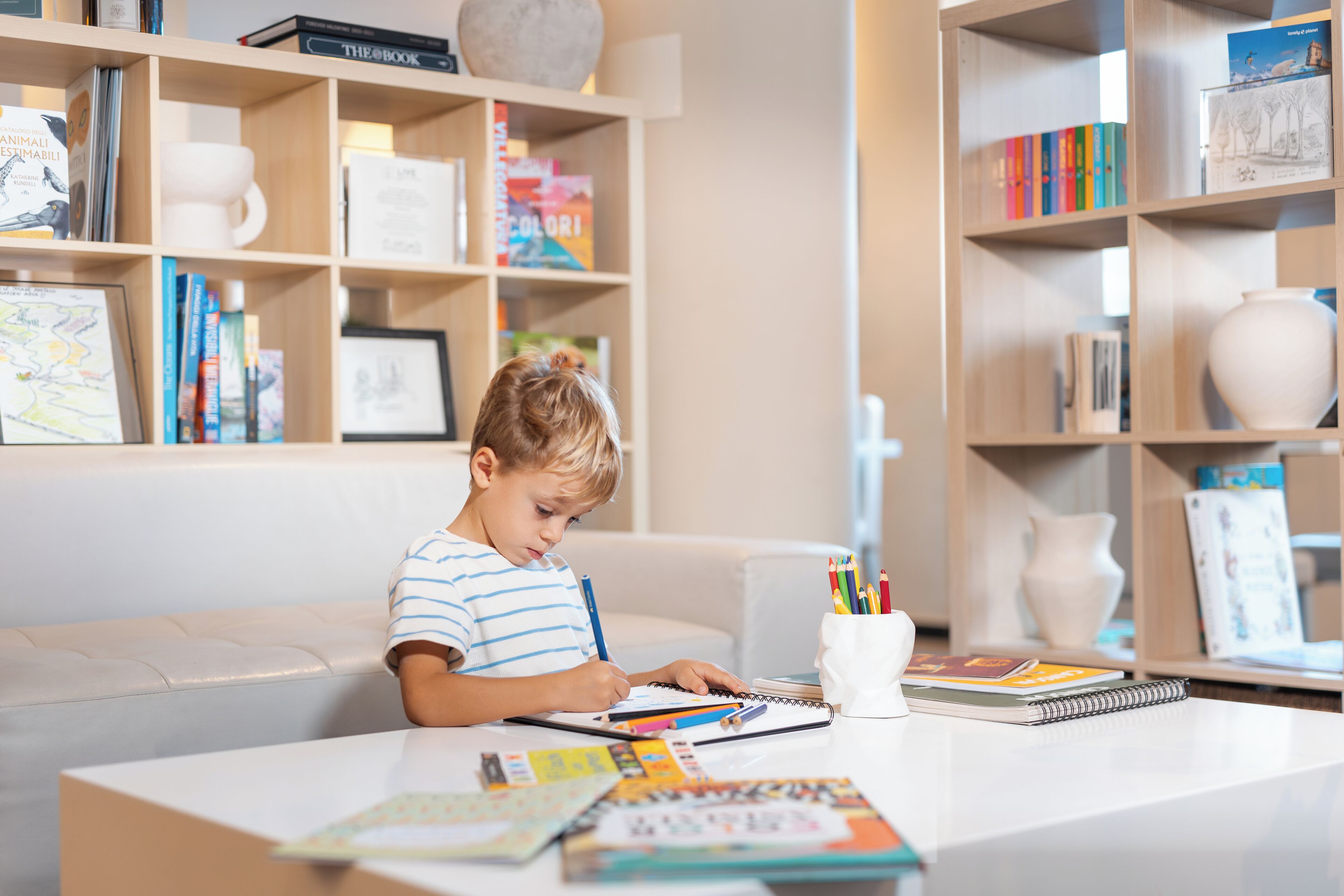 A child sitting at a table with books and art supplies, surrounded by shelves with books and decorative items.
