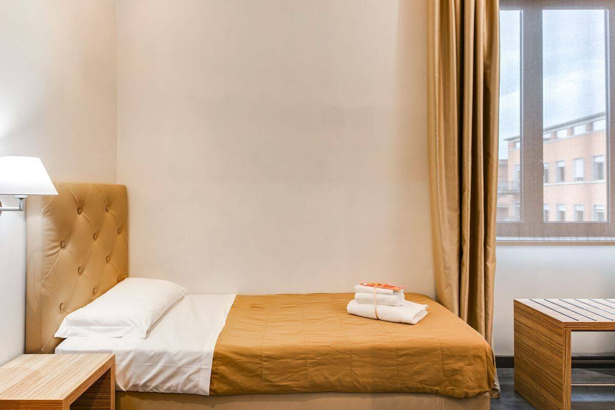A hotel room with a bed, a bedside table, a lamp, and a view of buildings through the window.