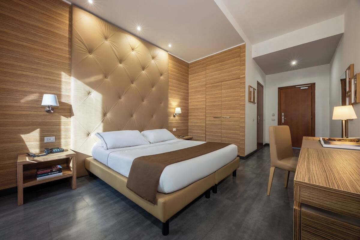 A modern hotel room with a large bed, wooden panel walls, a wooden desk, and a chair.