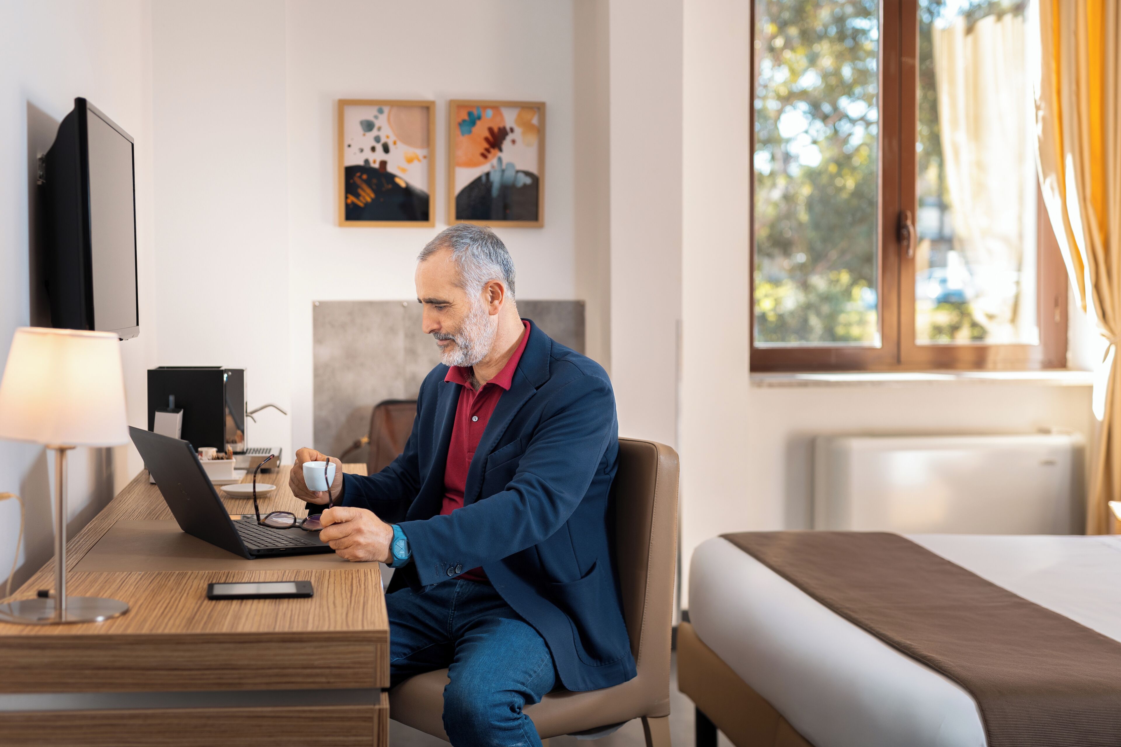 Un uomo seduto a una scrivania con un portatile, che tiene occhiali e una tazza, in una stanza con un letto, una televisione e opere d'arte alle pareti.