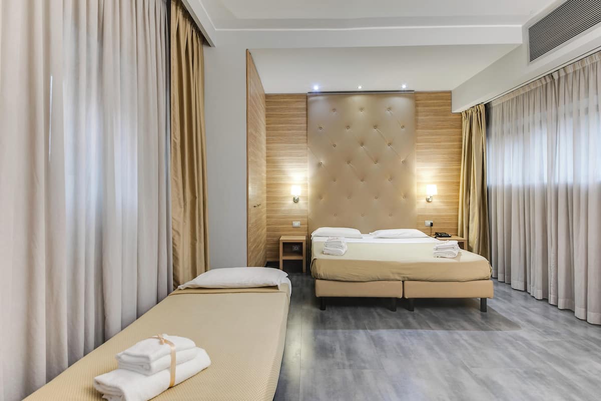 A hotel room with a large bed, a smaller bed, and a sofa bed. There is a wooden headboard, a nightstand, and a lamp.