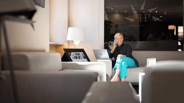 A person sitting on a white couch with a laptop and a framed photo, in a room with a large window and a wall-mounted lamp. - Phi Hotel Garden Area Roma Eur (Rome)