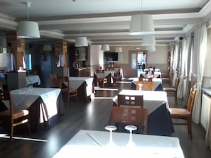 A spacious dining area with multiple tables and chairs, pendant lighting, and a view of the kitchen in the background. - Hotel Buenos Aires (Burgos)