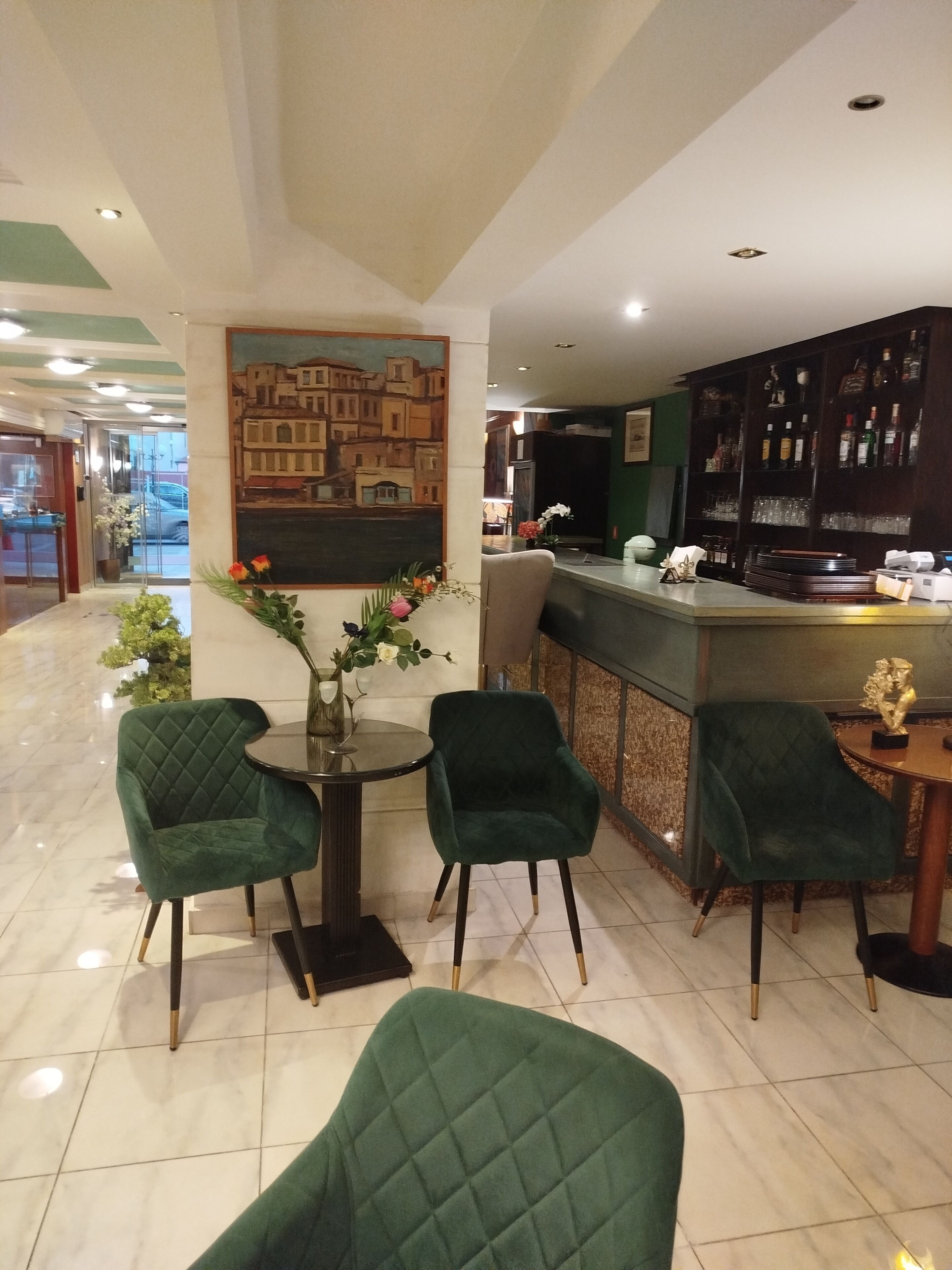 A hotel lobby with a bar area, green upholstered chairs, a round table with a vase of flowers, and a painting on the wall.