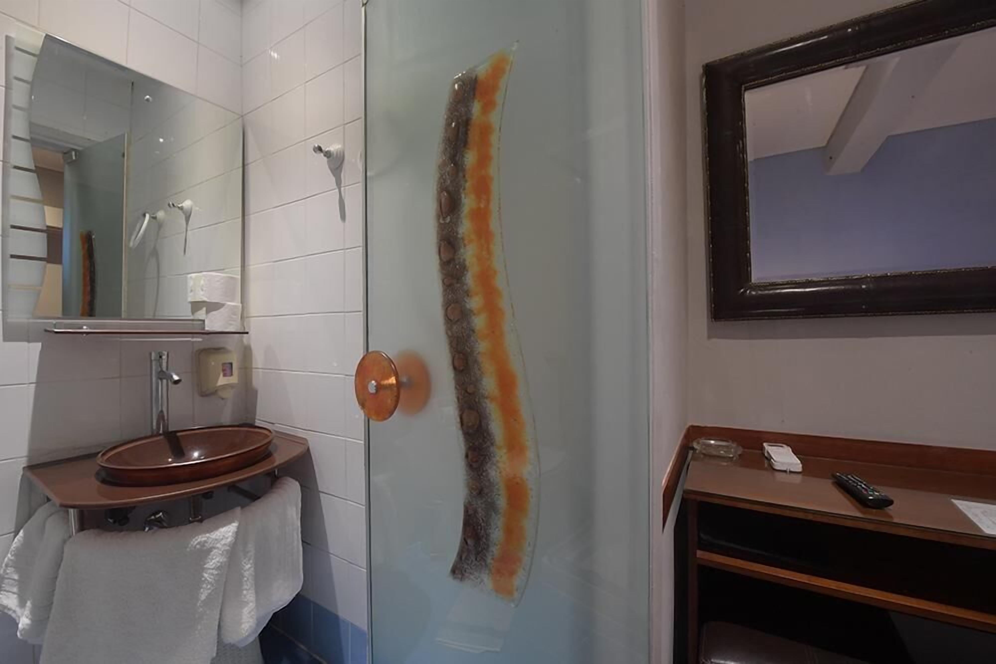 A bathroom with a glass shower door, a sink with a towel, a mirror, and a wooden vanity with a phone and remote control.