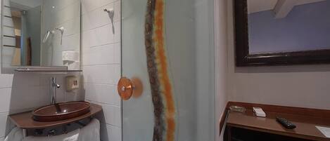 A bathroom with a glass shower door, a sink with a towel, a mirror, and a wooden vanity with a phone and remote control.
