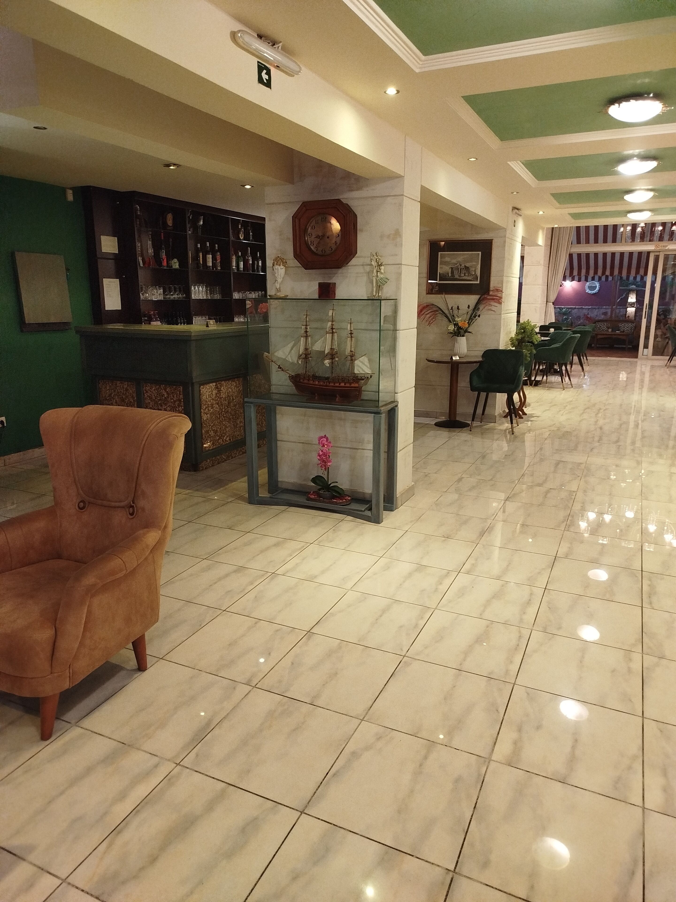 Un hall con zona de bar, una vitrina de cristal con un modelo de barco, una silla verde y un área de comedor con sillas verdes.