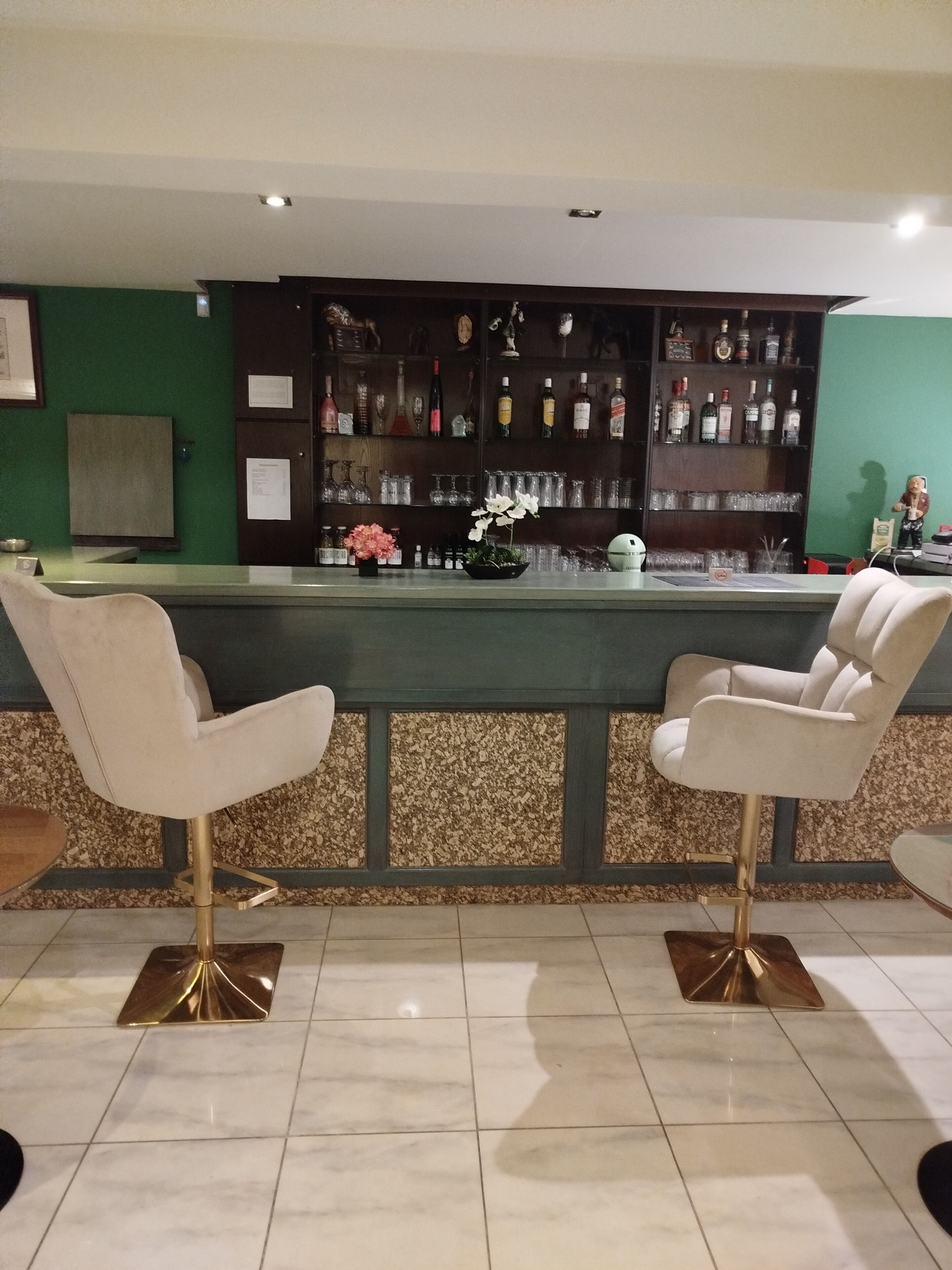 A bar area with two white chairs, a counter, and a green wall with shelves of bottles and glasses.