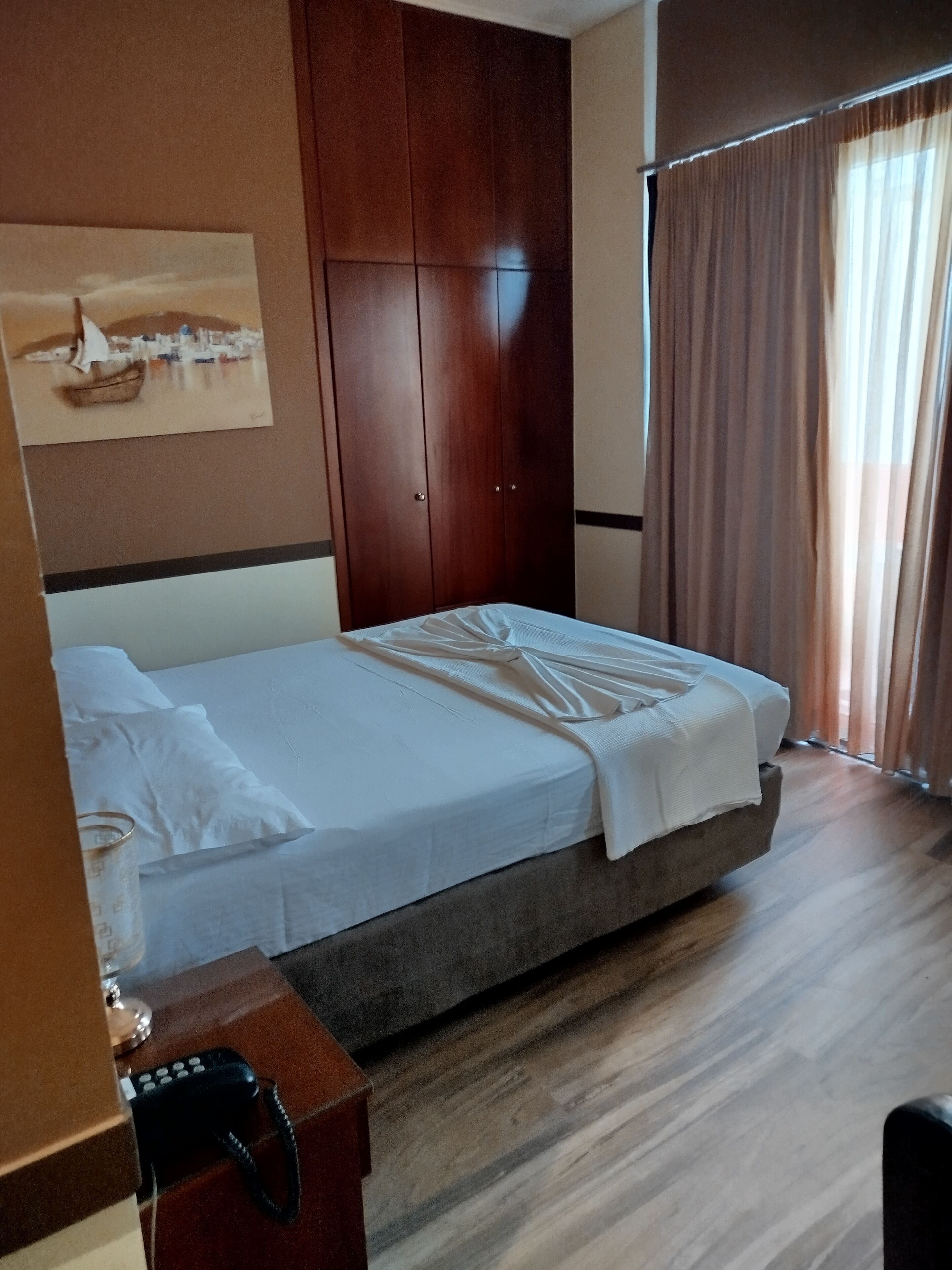 A hotel room with a bed, a wardrobe, a painting on the wall, and a desk with a telephone.