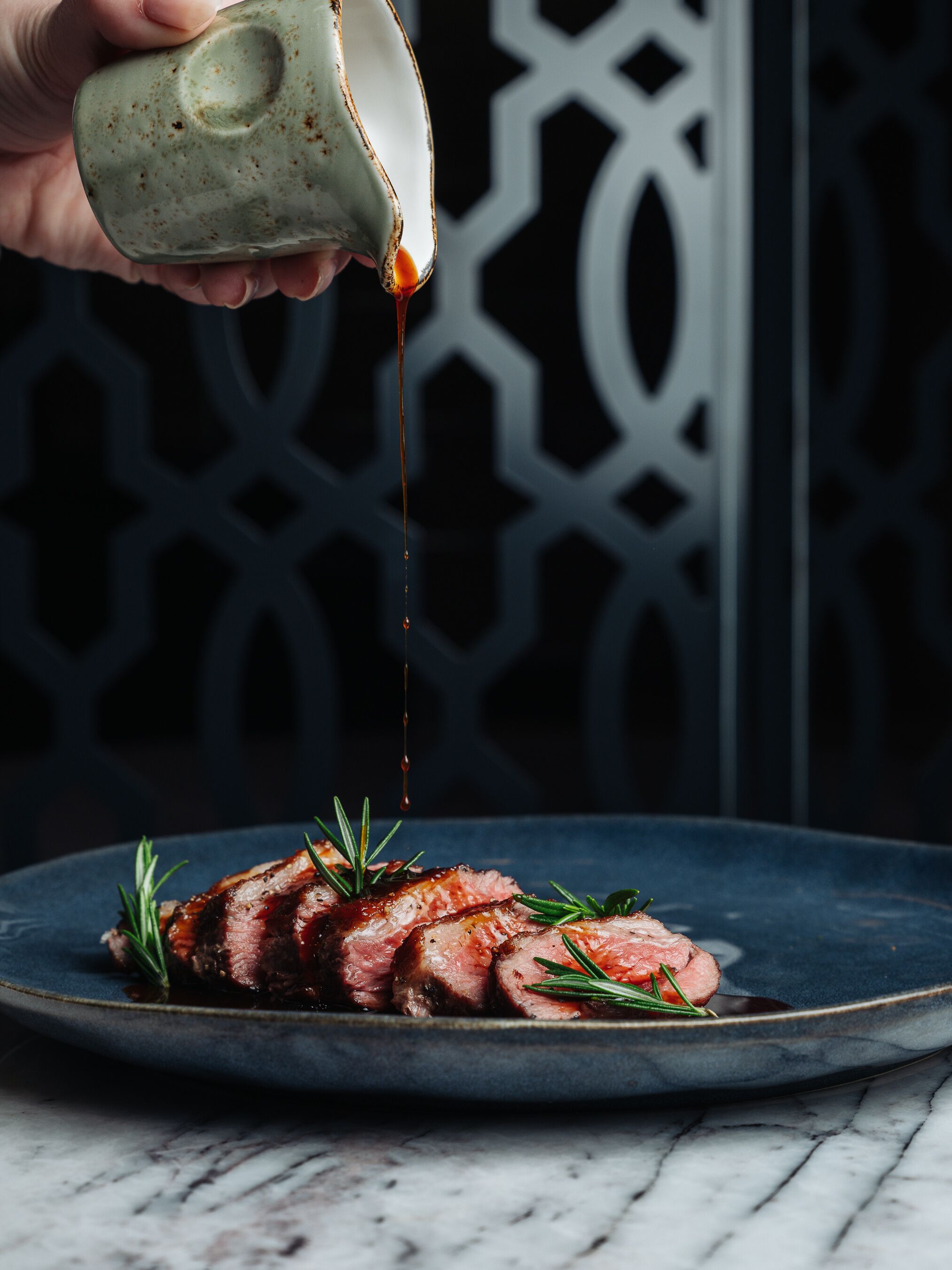 A plate of sliced meat garnished with rosemary, with a hand pouring sauce over it.