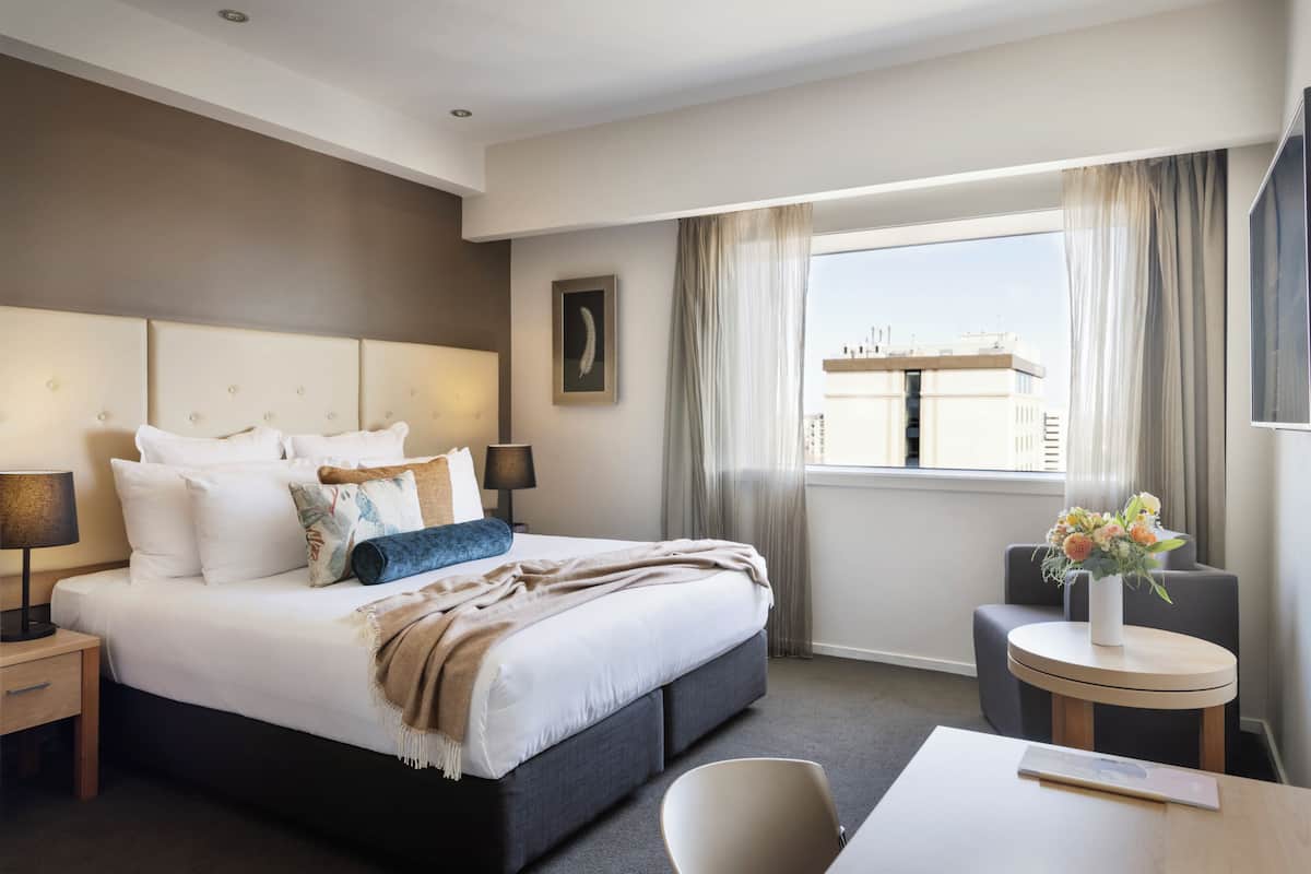A hotel room with a large bed, a bedside table, a chair, a small table, and a window with curtains.