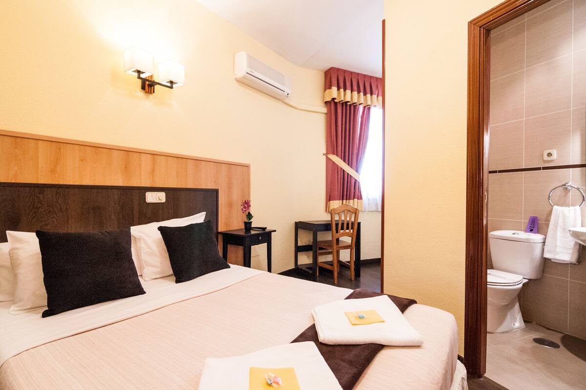 A hotel room with a large bed, a desk, a chair, a toilet, and an air conditioning unit.
