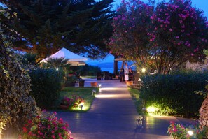A pathway leading to a covered outdoor seating area with tables and chairs, surrounded by lush greenery and flowering plants.