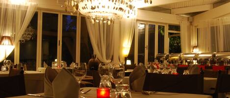 A dining area with tables set for a meal, a chandelier, and large windows.