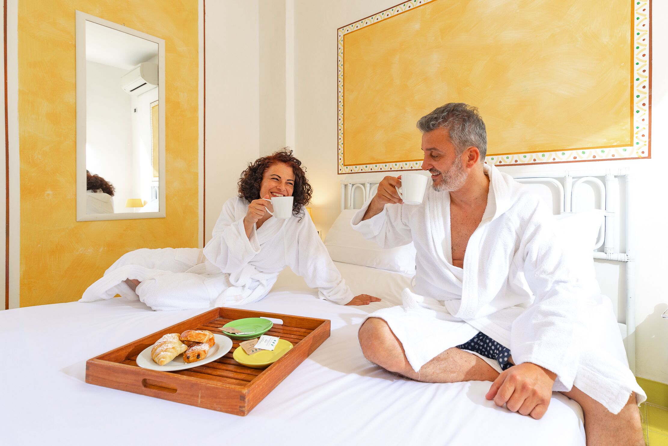 A couple in a hotel room with a bed, a tray with pastries, and a large yellow wall.