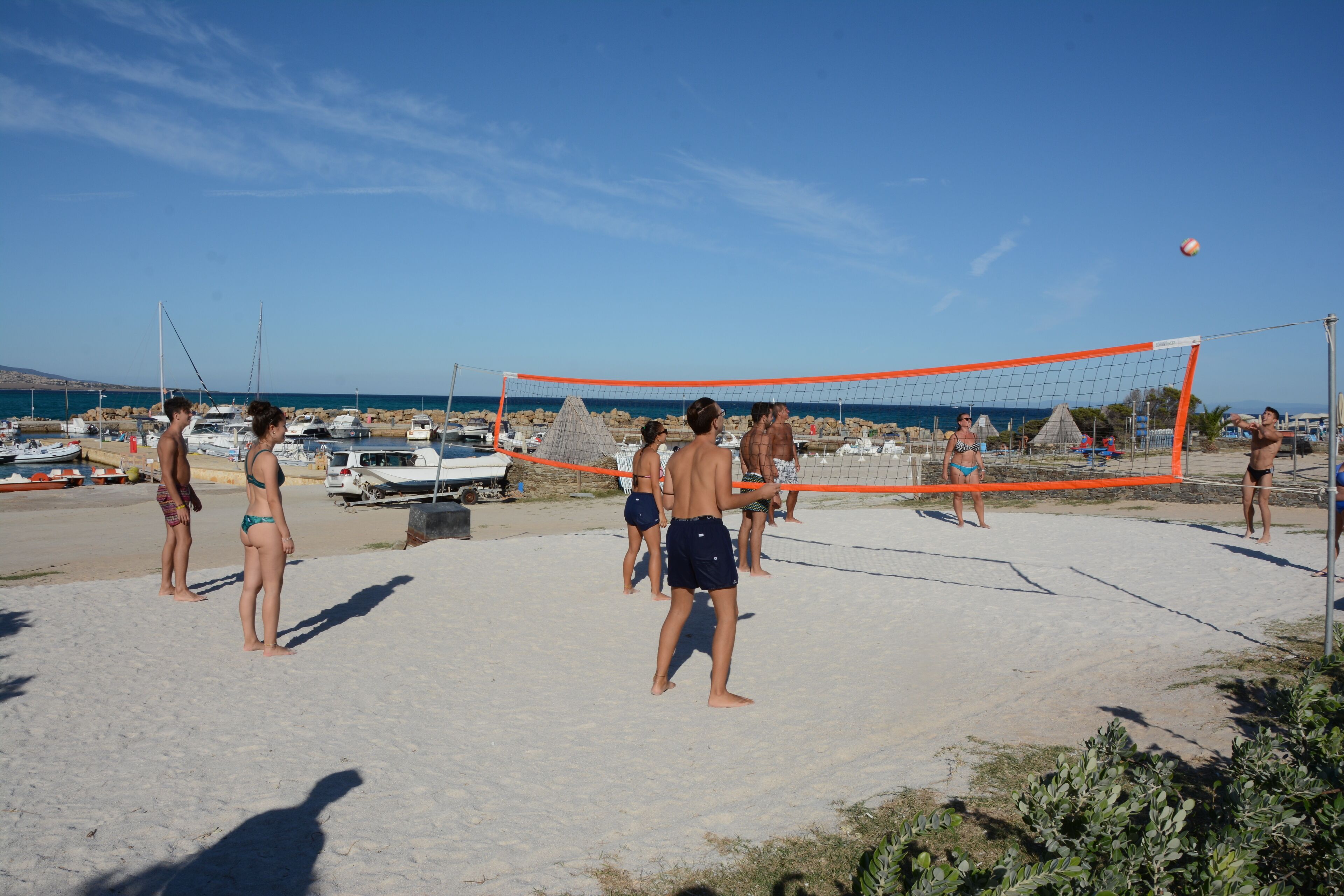 Partido de voleibol de playa con jugadores en traje de baño, una red y barcos al fondo.