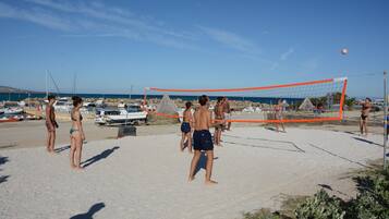 Partido de voleibol de playa con jugadores en traje de baño, una red y barcos al fondo.