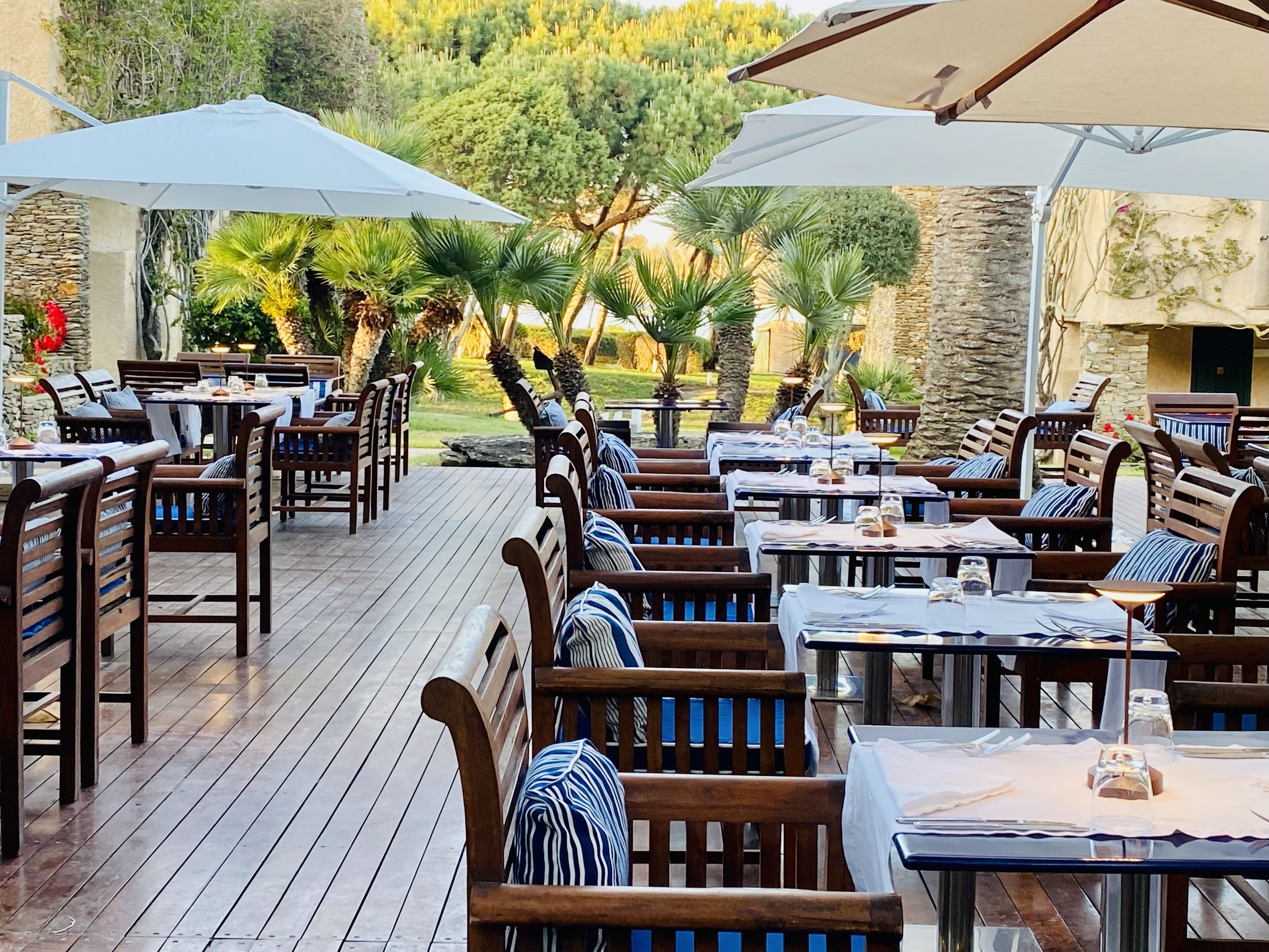 An outdoor dining area with wooden tables and chairs, covered by large umbrellas, set against a backdrop of greenery and stone walls.