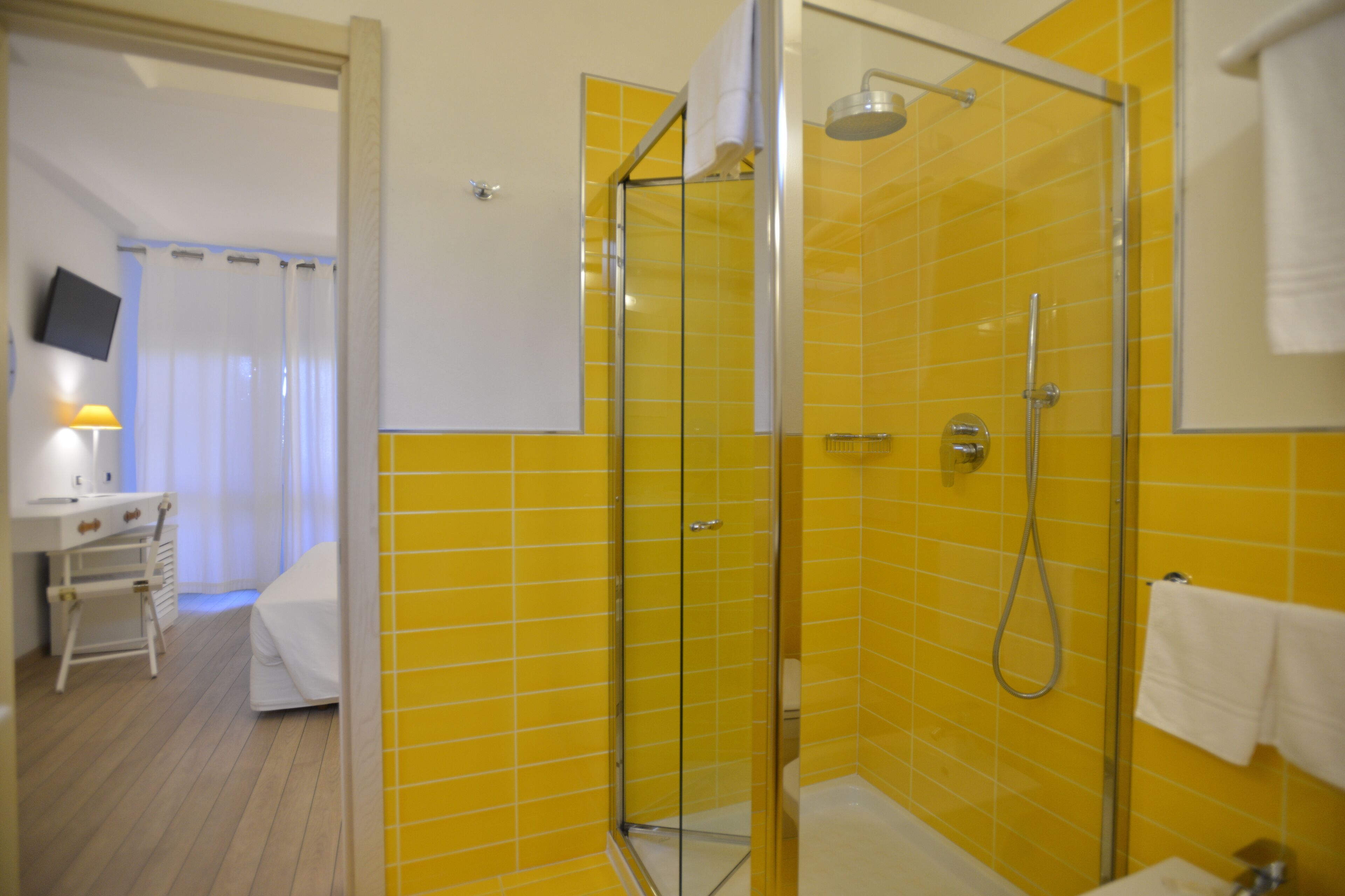 Un baño de hotel con baldosas amarillas, cabina de ducha de vidrio, lavamanos blanco y un televisor de pantalla plana sobre un escritorio.