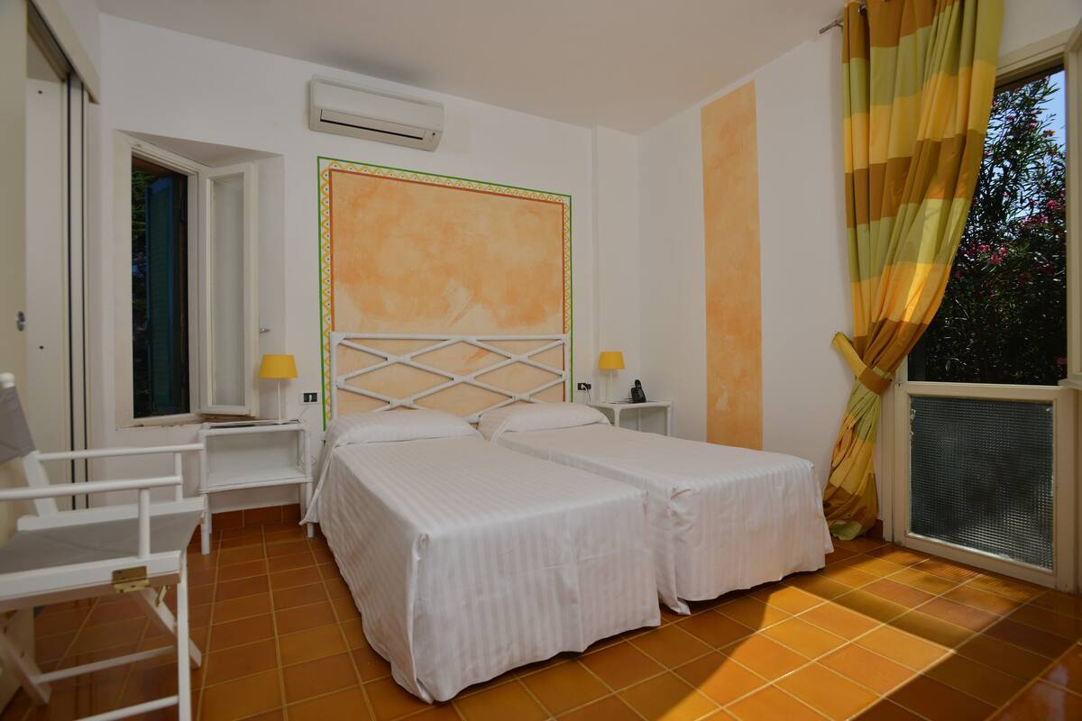 A room with two beds, a window with curtains, a small table with a lamp, and a white chair.