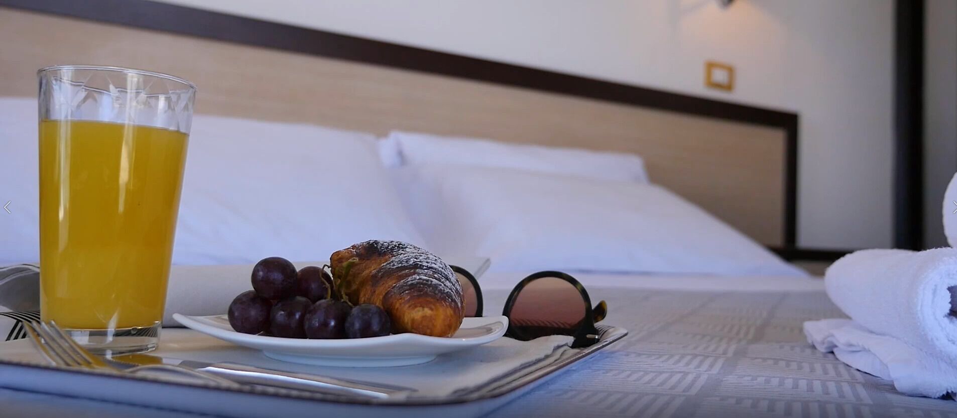 Une table de petit-déjeuner avec un croissant, des raisins et du jus d’orange sur un plateau, avec un lit et un oreiller en arrière-plan.