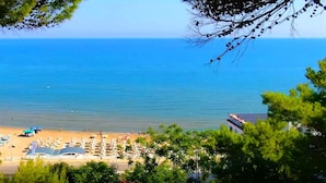 Ein Strand mit vielen Sonnenschirmen und Liegestühlen, freier Blick auf das Meer und ein mit 'HOTEL' beschriftetes Gebäude.