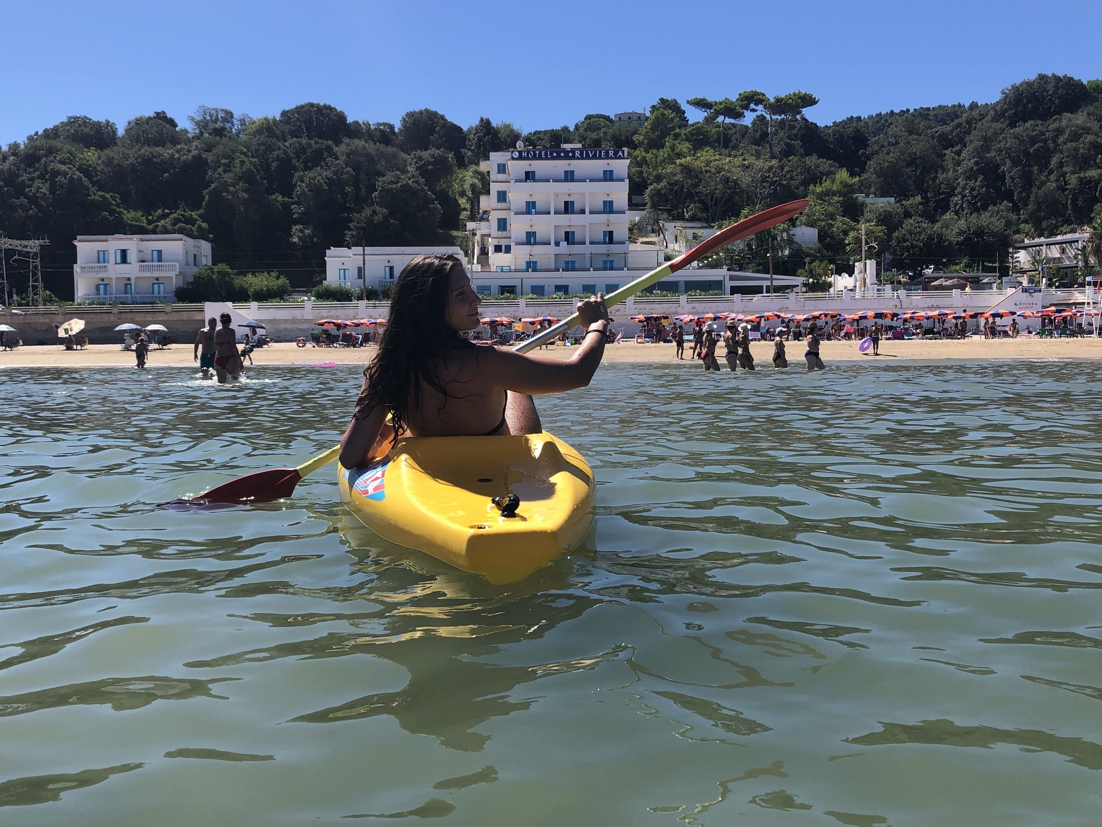 Een persoon in een gele kajak op het water, met een strand en een hotel op de achtergrond.