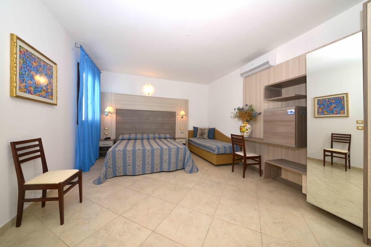 A bedroom with a bed, two chairs, a sofa, a bedside table, and a mirror.