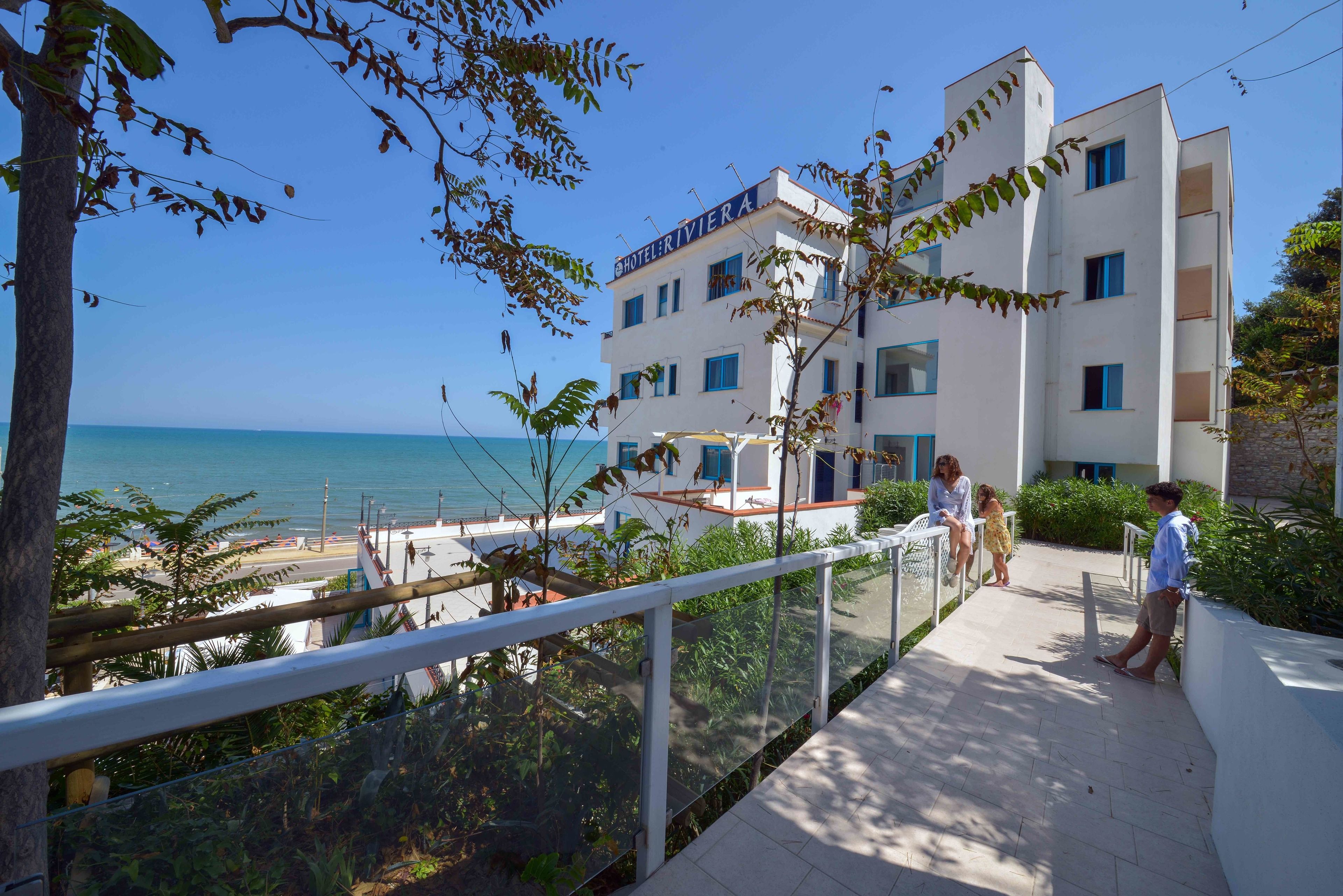A hotel with a balcony overlooking the sea, a clear sky, and a pathway with people.